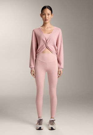 Vrouw die naar voren kijkt, gekleed in een mauve roze geribbelde trui, bijpassende sportbeha, leggings met hoge taille en witte sneakers met mauve veters.