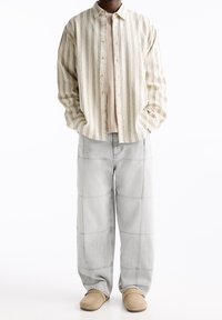 Chemise à rayures beige et grise, boutonnée, portée par-dessus un top beige uni, associée à un pantalon ample gris clair et des chaussures beige à enfiler.