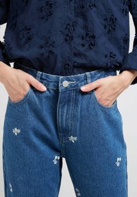 Jean en denim de couleur bleu moyen avec une broderie florale discrète. Design taille haute avec un bouton à l'avant et des poches latérales.