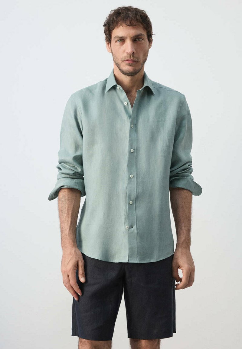 Homme portant une chemise boutonnée vert clair à manches retroussées et un short foncé au niveau des genoux, debout devant un fond uni.