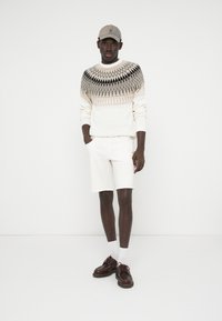 Strickpullover mit geometrischem Muster in Creme, Beige und Schwarz; weiße Shorts; braune Schuhe; graue Mütze; einfarbiger Hintergrund.