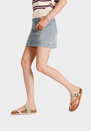 Donna con minigonna in denim, top a righe e sandali bicolore con cinturino e fibbia che cammina su uno sfondo semplice.