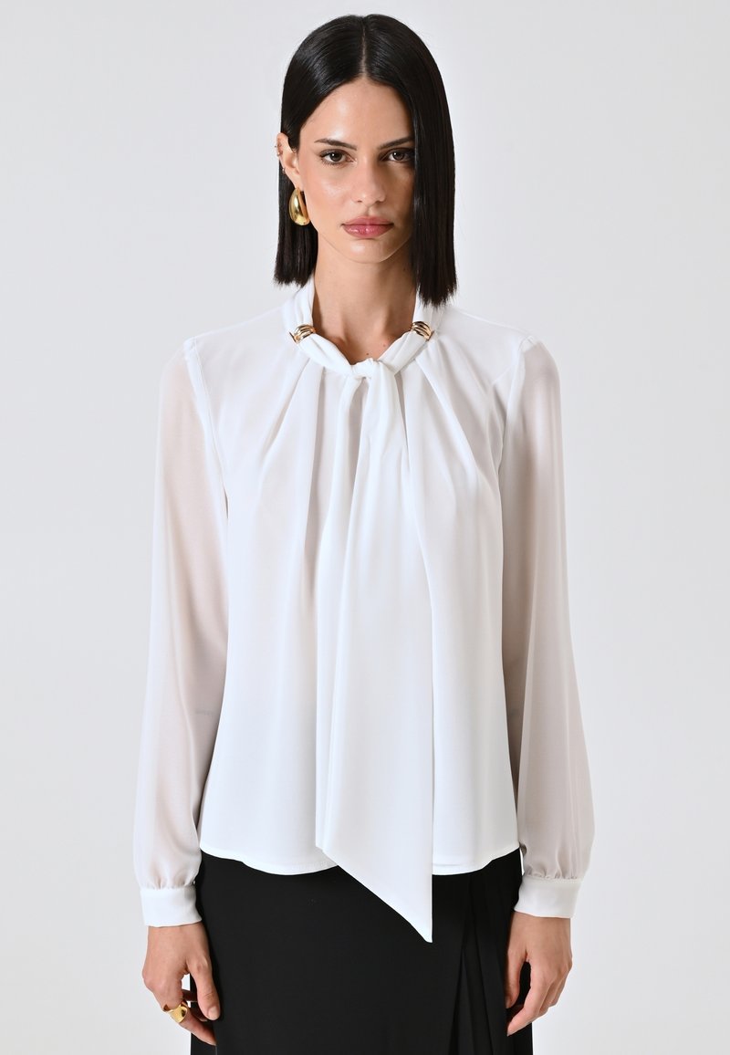 Blusa bianca realizzata in tessuto leggero con un design drappeggiato, dotata di dettagli in metallo dorato al collo, maniche lunghe e un pannello anteriore morbido.