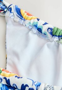 Góra bikini o białej bazie, ozdobiona kolorowymi wzorami kwiatowymi i spiralnymi, z marszczeniami na brzegach, wykonana z gładkiej tkaniny.