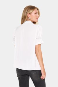 Blusa bianca a maniche corte con una morbida texture lucida. Presenta maniche a sbuffo e un'orlo leggermente curvato, abbinata a jeans scuri.