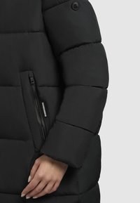 Schwarze Steppjacke mit einem gepolsterten Design, ausgestattet mit einer seitlichen Reißverschlusstasche, weichem Material und einem runden Logo auf der Schulter.