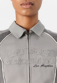 Giacca grigia chiara con zip frontale, dotata di colletto, ricamo "GUESS ORIGINALS" e piping bianco lungo le cuciture.