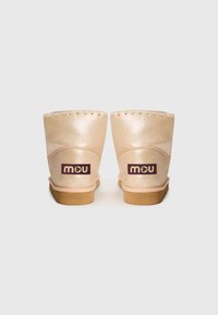 Béžové semišové kotníkové boty s lesklým povrchem, dekoračním švem kolem horní části a fialovým logem "mou" na zadní straně. Plná gumová podrážka.