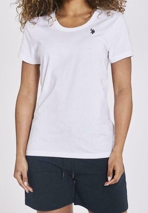 Femme portant un t-shirt blanc uni à manches courtes avec un petit logo noir sur la poitrine et un short foncé, debout, bras détendus.