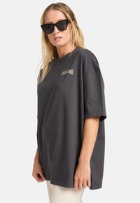 Overdimensioneret grå bomulds t-shirt med korte ærmer, rund halsudskæring og et lille logo foran. Bæres med tykt rammede sorte solbriller.