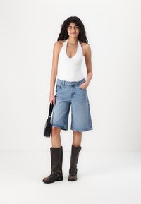Balta haltera tops, zili džinsa bermudu šorti un ceļgalu garuma brūni ādas zābaki. Modelis tur mazu melnu rokassomu. Vienkāršs fons.