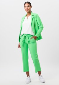 Heldergroene pak met een blazer en een broek. De blazer heeft een knoopdetail, de broek heeft een ceintuur in de taille, gecombineerd met een witte top en witte sneakers.
