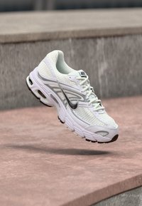 Bílé a šedé běžecké boty Nike s síťovinou a černou podrážkou plovoucí nad betonovým povrchem.