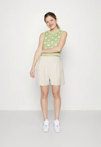 Zaļš teksturēts īss džempera tops un bēši šorti ar elastīgu jostasvietu. Modelis valkā baltas sporta apavi. Vienkāršs, ikdienišķs tērps.