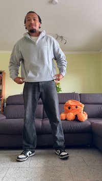 Corta-vento cinza com fecho e bolso à frente, jeans pretos e ténis pretos com detalhes brancos. Brinquedo de polvo laranja macio em sofá cinza.