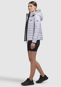 Lichtgrijze pufferjack met een zilveren capuchon, een rits aan de voorkant en logo-accenten, gecombineerd met zwarte shorts en sport schoenen.