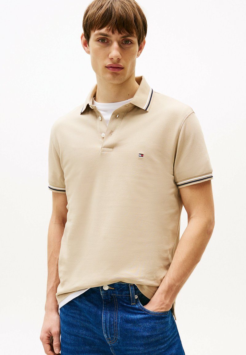 Beżowa koszulka polo z teksturowanej tkaniny, z trzema guzikami oraz kontrastującymi granatowymi paskami na kołnierzu i rękawach; małe logo na piersi.