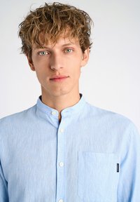 Lichtblauw overhemd met opstaande kraag, van textuurstof, met een enkele borstzak en een klein zwart etiket aan de zijkant.