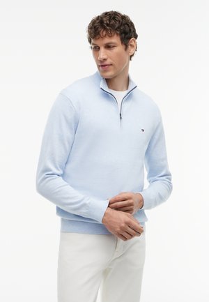 Homme aux cheveux bouclés portant un pull bleu clair à fermeture éclair sur le haut du torse et un pantalon blanc, regardant vers le bas les mains jointes devant lui.