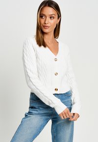 Cardigan cropped en tricot blanc avec un décolleté en V profond, doté de boutons en bois et de poignets côtelés, associé à un jean bleu clair.