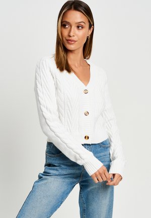 Cardigan cropped en tricot blanc avec un décolleté en V profond, doté de boutons en bois et de poignets côtelés, associé à un jean bleu clair.