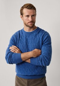 Suéter de punto azul con cuello redondo, puños y dobladillo acanalados, que presenta patrones texturizados. Lleva puesto un camiseta blanca debajo y se combina con pantalones color beige.