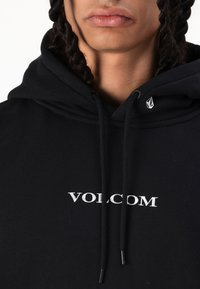 Fekete pamut kapucnis pulóver, kenguru zsebbel, fehér "VOLCOM" logóval és egy kis ezüst emblémával a bal vállen.