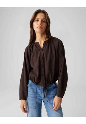 Frau trägt eine dunkelbraune, geknöpfte Bluse mit langen Ärmeln und blaue, hoch taillierte Jeans, steht vor einem schlichten, hellen Hintergrund.