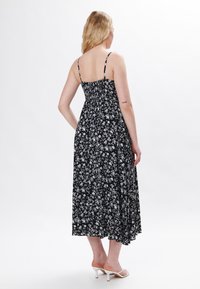 Schwarzes Maxikleid mit floralen Mustern, dünnen verstellbaren Trägern, einem gerafften Rückenteil und einem fließenden Design. Es zeigt weiße Blumenmuster auf einem dunklen Hintergrund.