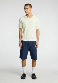 Polo shirt bianco a righe con maniche corte e colletto, abbinato a pantaloni corti blu navy a ginocchio, calze bianche e mocassini neri.