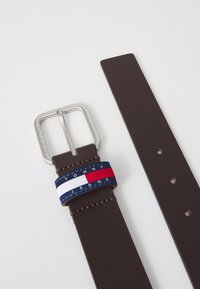 Brun lærrem med en sølvfarget spenne, prydet med et bånd som har røde, hvite og blå striper og teksten "TOMMY JEANS".