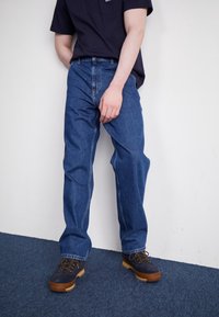 Siniset farkut suoralla lahkeen leikkauksella, joissa näkyvät saumat ja perinteinen nappisuljenta. Yhdistetty tummiin, nauhakiinnitteisiin kenkiin.