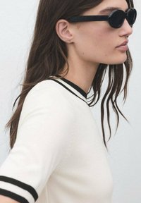 Woman with long brown hair wearing black oval sunglasses and a white shirt with black trim on collar and sleeves, facing right.