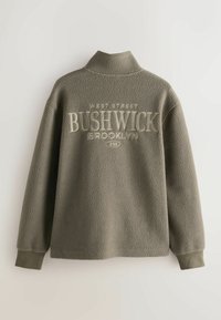 Polarowy pulower w oliwkowej zieleni z wysokim kołnierzem, z napisem "BUSHWICK BROOKLYN" w kremowym kolorze, teksturowana powierzchnia oraz prążkowane mankiety.