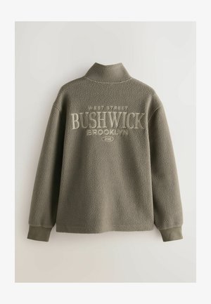 Fleece-pull med hög krage i olivgrön färg, med texten "BUSHWICK BROOKLYN" i krämvit, texturerad yta och ribbade ärmslut.