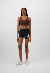 Un soutien-gorge de sport bordeaux et un short noir avec un logo Nike blanc. Les baskets sont de couleur rose clair. Le matériau semble léger, adapté aux vêtements de sport.