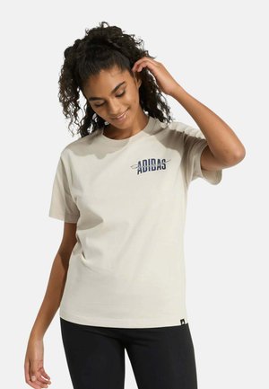 Mujer con cabello rizado que lleva una camiseta beige de Adidas y pantalones negros, sonriendo y tocándose el cabello sobre un fondo liso.