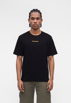 Bărbat cu părul împletit, purtând un tricou negru cu text galben mic "Saint-Tropez" și pantaloni scurți cargo olive, stând în fața unui fundal simplu.