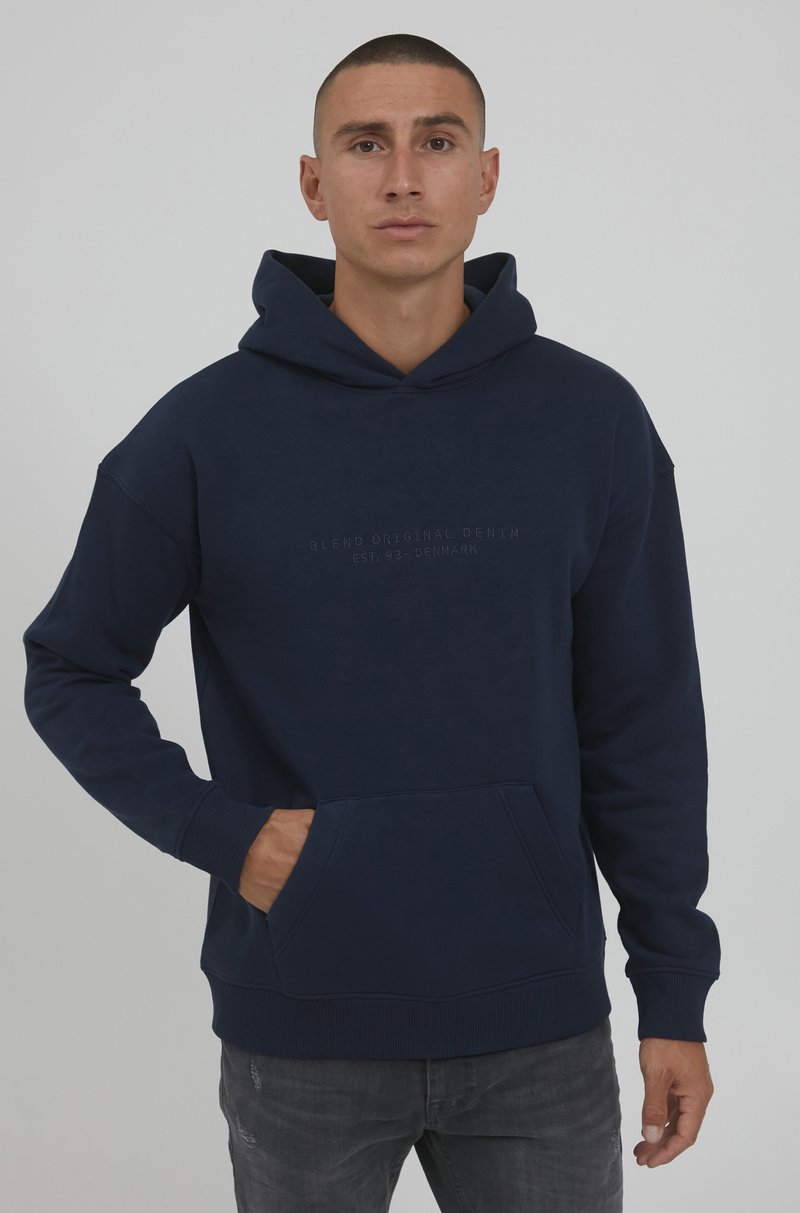 Navy blue hooded sweatshirt made of soft fabric; features a front kangaroo pocket and embossed branding on the chest. Boxy fit and ribbed cuffs.