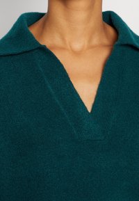 Detailní záběr na krk a horní část hrudníku osoby, která nosí světle modrý pletený svetr s širokým V-výstřihem.