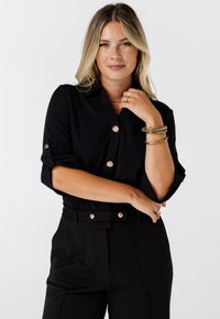 Blonde vrouw met lichte make-up draagt een zwarte blouse met knopen en een broek, geaccentueerd met gouden creolen, armbanden en ringen, poseert zelfverzekerd.