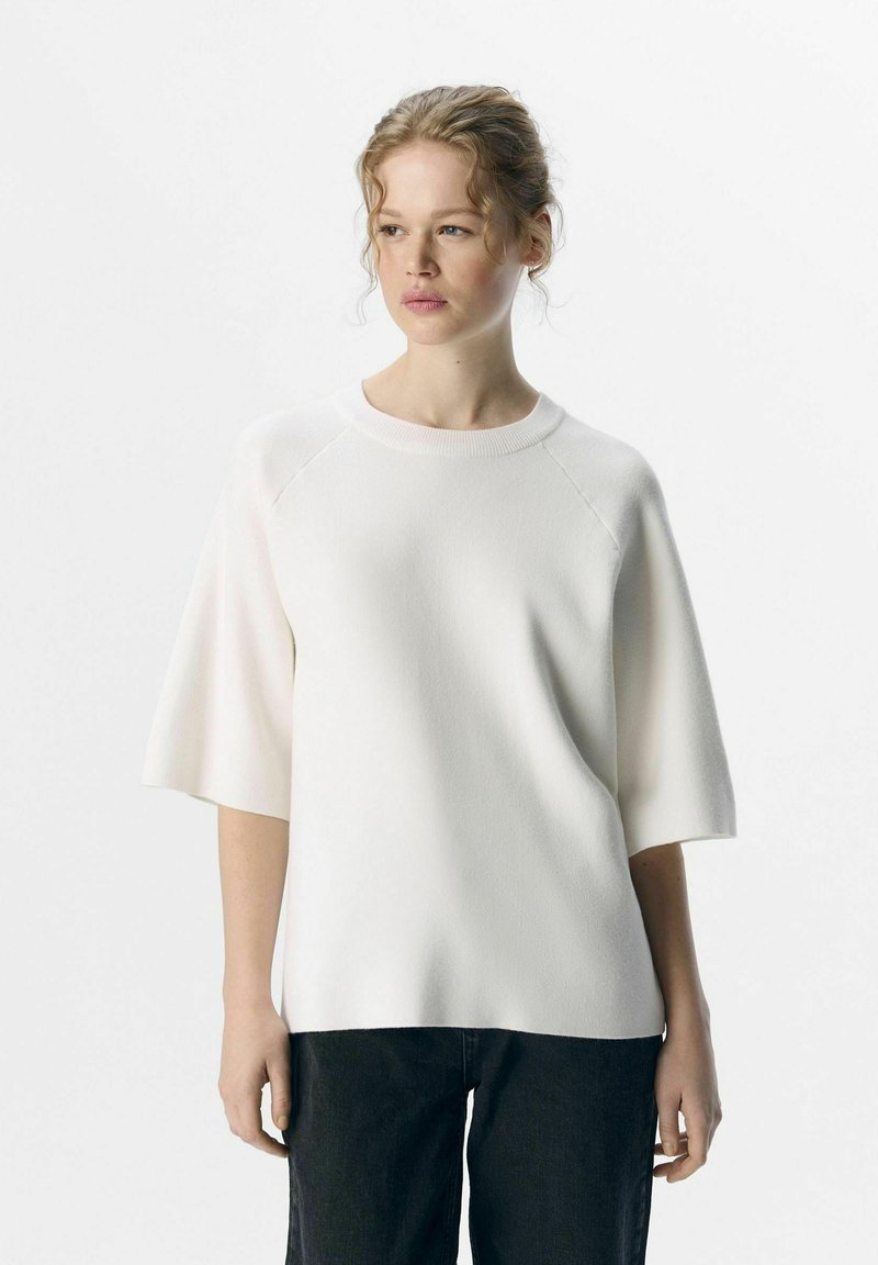 Haut blanc à manches courtes avec un col rond, fabriqué en tissu texturé, présentant des manches raglan et une coupe décontractée, associé à un jean foncé.