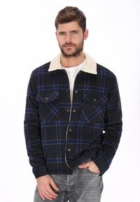 Chaqueta de franela a cuadros en azul marino con detalles rojos, con un cuello de piel sintética color crema, dos bolsillos en el pecho y cierre de botones.