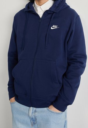 Person trägt einen marineblauen Nike-Zip-Hoodie mit Taschen über einem weißen Hemd und hellblauen Jeans, die Hände in den Taschen.