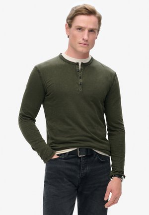 Grøn langærmet Henley-trøje med en to-knaps placket, lagd over en lys T-shirt, parret med mørke jeans og et sort bælte.