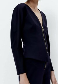 Blusa blu navy con zip, scollo a V, maniche lunghe e dettagli in oro. Presenta un design aderente e una discreta lavorazione dei cuciture.