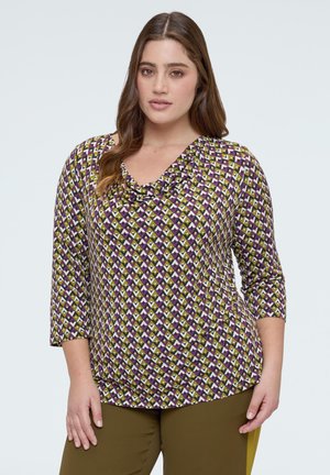 Mujer con cabello largo y castaño, vestida con una blusa de patrón en tonos morado, blanco y oliva con mangas tres cuartos y pantalones color oliva, de pie contra un fondo liso.