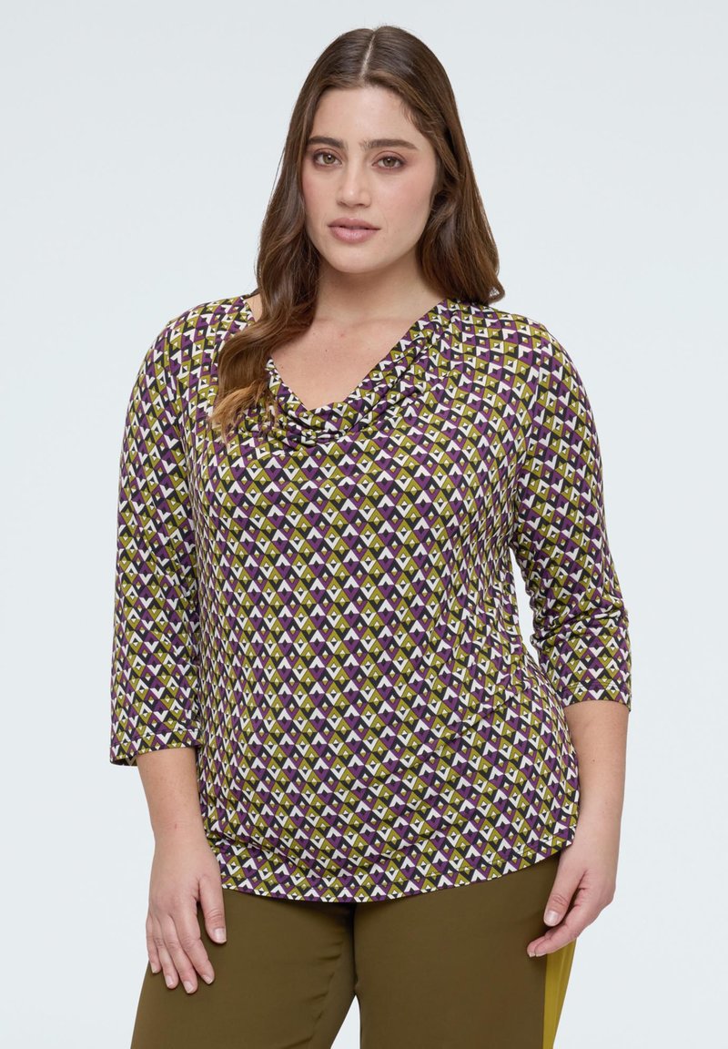 Femeie cu păr lung și castaniu, purtând un top cu mâneci trei sferturi, cu model în nuanțe de mov, alb și măsliniu, și pantaloni măslinii, stând pe un fundal simplu.