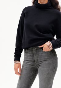 Žena v černé mikině s rolákem, šedých džínách s vysokým pasem a proužkovaném tričku s dlouhým rukávem pod ní, s rukou v kapse.