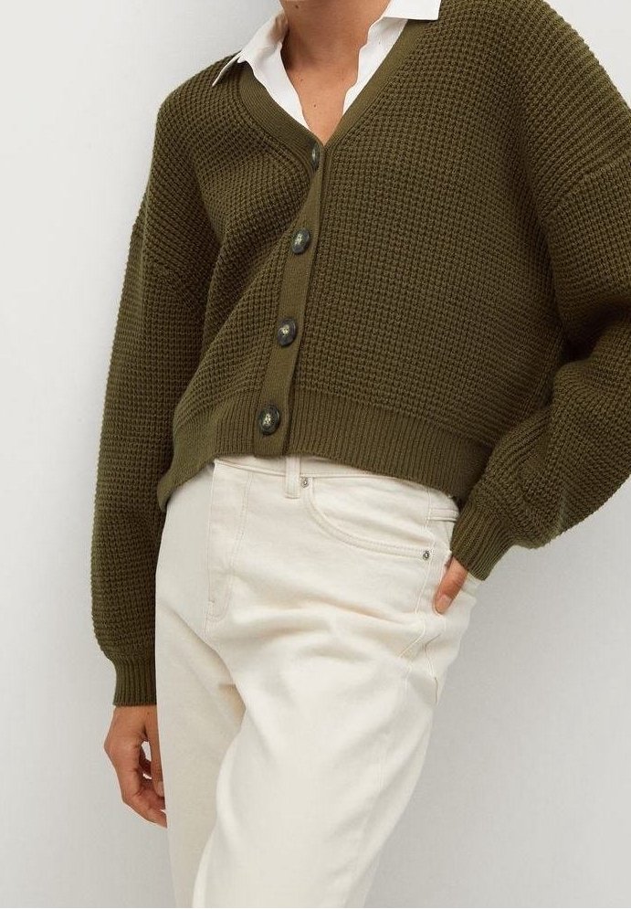 Personne portant un cardigan boutonné texturé vert olive sur une chemise blanche et un pantalon écru, une main dans la poche, fond neutre.